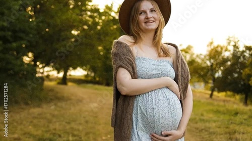 Smiling pregnant woman in summer dress and hat gently holding her belly while standing outdoors during golden hour. Concept of maternity lifestyle, pregnancy health, wellness, and emotional wellbeing
