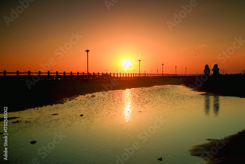 A breathtaking sunset at Yong-an Fishing Harbor, Taiwan, casting a warm orange glow across the sky. Silhouetted figures add a sense of human presence to the serene winter scene.