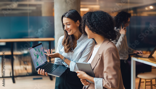 In a bright, modern office setting, two women engage in a lively discussion while working on a laptop together