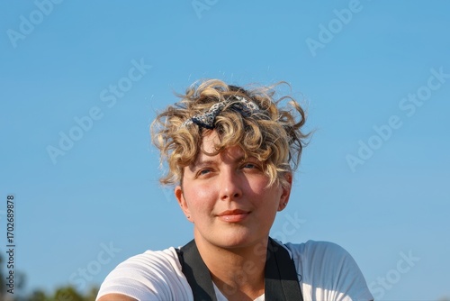 Close up portrait head shot of  natural looking beautiful middle aged Caucasian woman with short curly blond hair outdoors outside looking off into the distance in nature with blue sky background