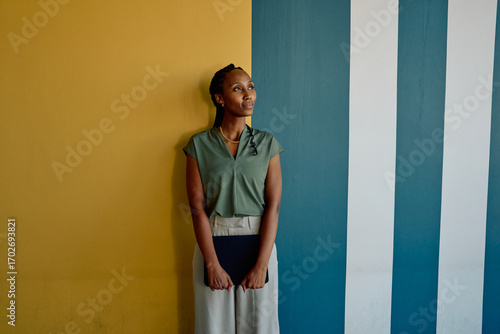 African young businesswoman with tablet looking up while standing against colorful wall in office