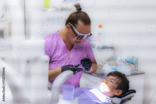 A dentist in purple scrubs examines a young boys teeth while he sits in the dental chair, wearing protective glasses and gloves during the procedure.