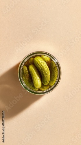 Top-down view of whole pickles packed in a glass jar, placed on a soft beige surface with long shadows.