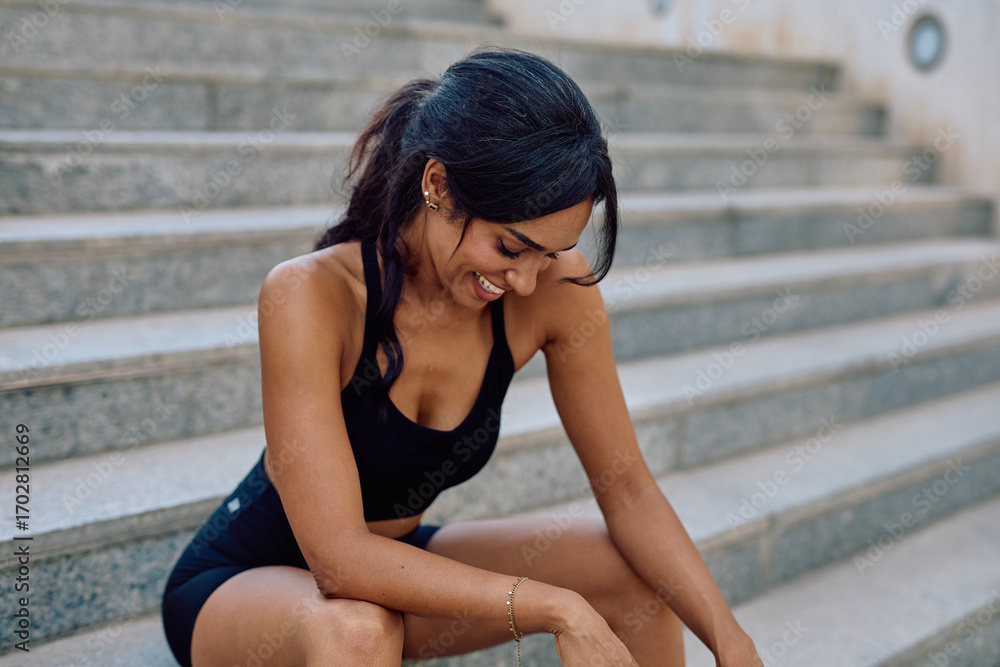 Obraz premium Young woman resting on urban stairs, smiling and enjoying a moment of relaxation after an invigorating training session in sportswear