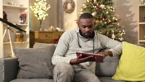 Handsome African guy sitting on couch and holding digital tablet in hands. Young man searching something in internet. Indoors. Christmas atmosphere.