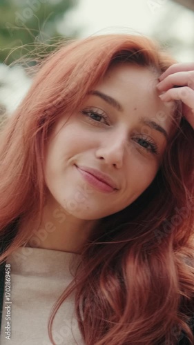Vertical video, Beautiful redhead woman smiles and looks at camera. She's standing in a park and runs her hand through her hair. Medium shot.
