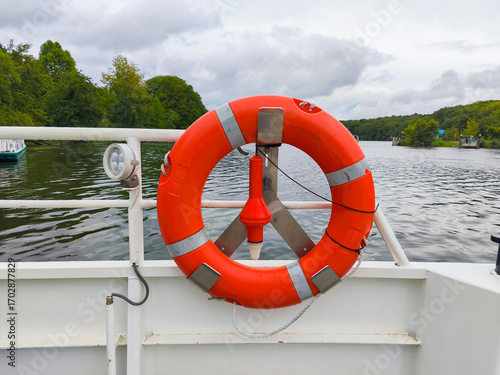 Berlin, Germany - September 05, 2025: Bright orange life buoy mounted on a boat railing, surrounded by tranquil water and lush green trees, emphasizing safety on the water