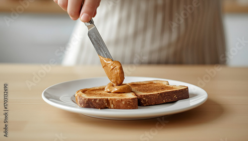 In the foreground is a white plate with two slices of bread on which peanut butter is applied. A hand holds a knife, spreading peanut butter on bread.