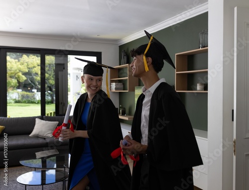 Celebrating graduation, teens in gowns smiling and holding diplomas at home