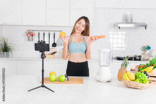Happy young asian woman in blue activewear create recipe video content fresh fruit and vegetable for healthy smoothie recipe in her bright kitchen holding a carrot and orange.