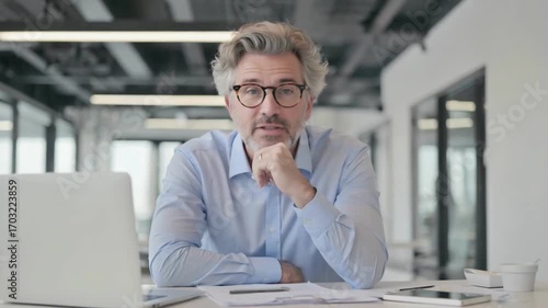 Thoughtful Reflection: A focused individual in eyeglasses, deeply contemplating, possibly on professional matters, as a laptop and office documents lie at the ready.