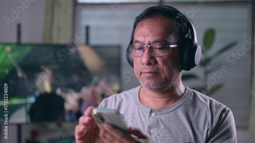 Aged person enjoying digital media, wearing comfortable headphones, intently watching phone screen content. Relaxed human engaging modern tech, gaming, streaming media home. Leisure pursuit, personal