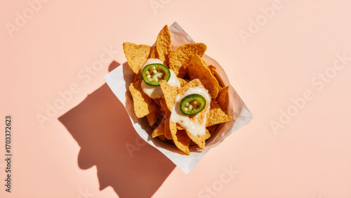 Nachos in a plain kraft boat with melted cheese and jalapeño slices on a pale peach background, slight overhead studio shot with soft contact shadows
