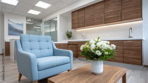 A modern office interior featuring a light blue armchair, wooden cabinets, and a flower arrangement on a table, creating a welcoming and stylish workspace.