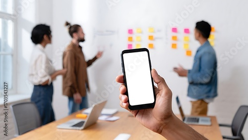 A person holds a smartphone with a blank white screen during a creative team brainstorming meeting in an office.