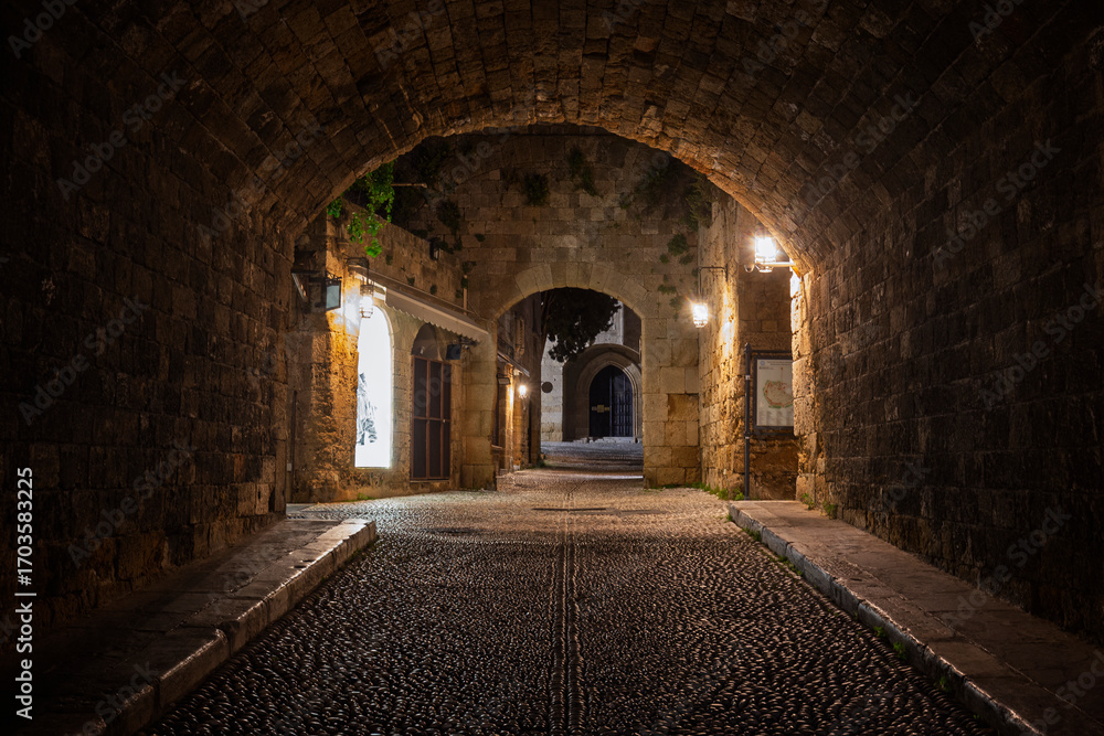 Fototapeta premium Idyllic and empty cobblestone street and tunnel at the medieval UNESCO World Heritage Site of Old Town of Rhodes on Rhodes island in Greece at night.