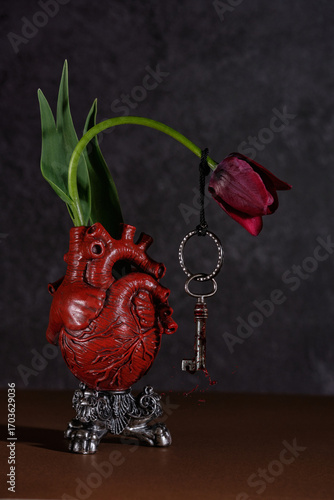 Modern still life, frikibana. Red vase in the shape of an anatomical heart with a red tulip in it. The tulip bends under the weight of the key hanging on it. The key is splashed with red liquid, blood