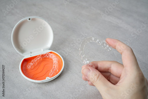Clear dental aligner in hand and orange case on table. Dental orthodontic mouthpiece.