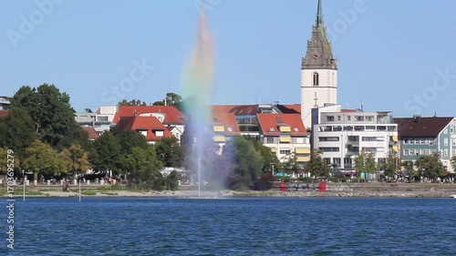A scenic view of a lakeside fountain and a church tower in Friedrichshafen, Germany, under a clear blue sky on a sunny summer day