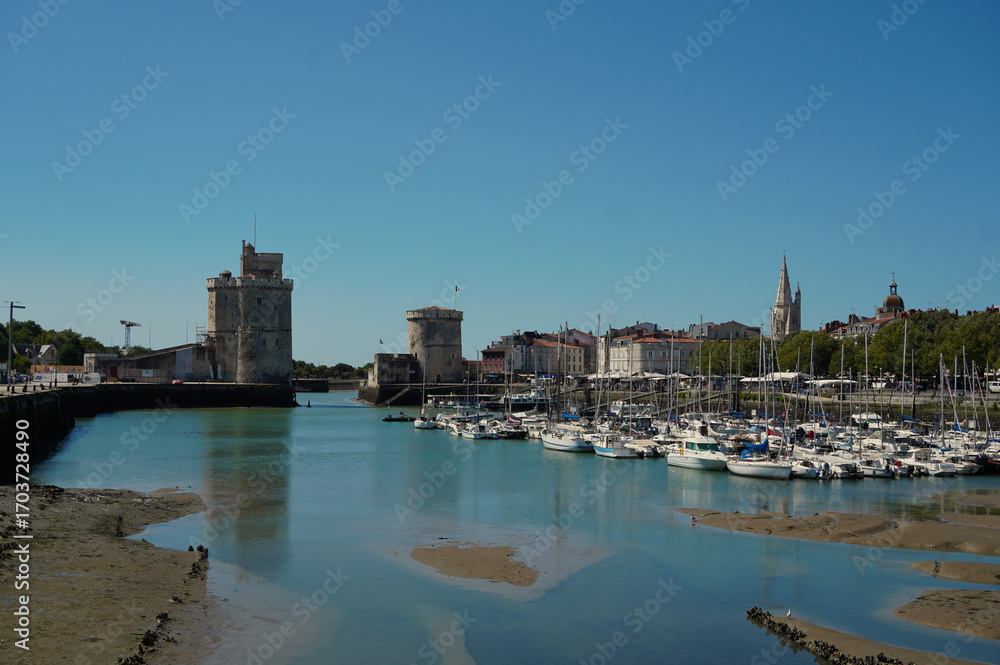 Naklejka premium La Rochelle marina with sailboats and boats docked. In the background, the iconic medieval towers guarding the entrance to the historic French port. Clear blue sky overhead.