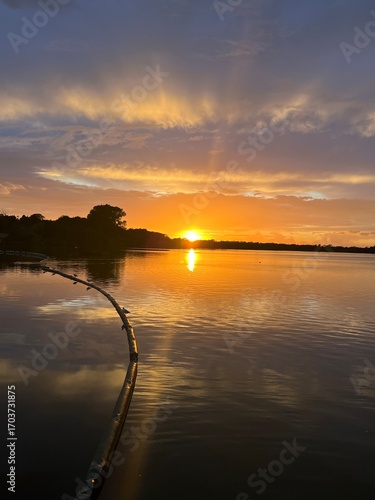 Summer sunset landscape sun setting over Norfolk Broads England uk tranquil water with the beautiful orange yellow blue skies viewed from sailing boat to horizon and trees reeds, clouds reflected 