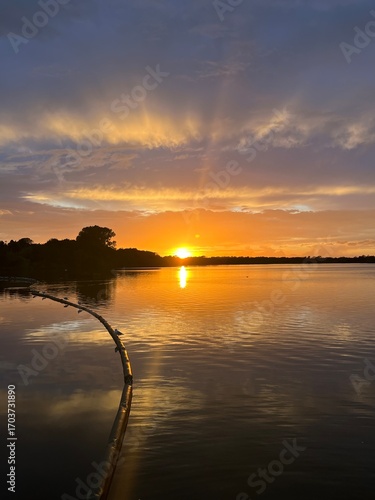 Summer sunset landscape sun setting over Norfolk Broads England uk tranquil water with the beautiful orange yellow blue skies viewed from sailing boat to horizon and trees reeds, clouds reflected 