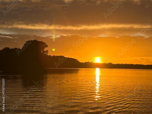 Summer sunset landscape sun setting over Norfolk Broads England uk tranquil water with the beautiful orange yellow blue skies viewed from sailing boat to horizon and trees reeds, clouds reflected 
