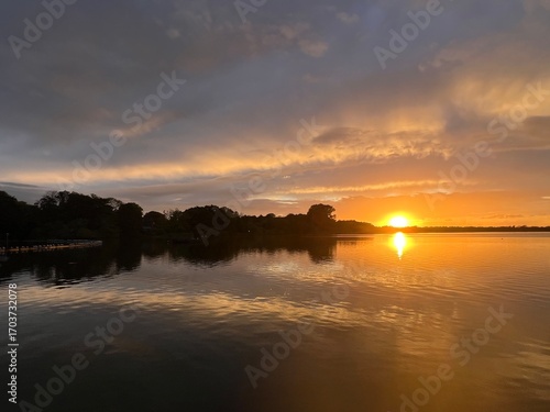 Summer sunset landscape sun setting over Norfolk Broads England uk tranquil water with the beautiful orange yellow blue skies viewed from sailing boat to horizon and trees reeds, clouds reflected 