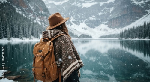 A woman in a tan hat and poncho stands by a snowy winter lake, gazing at the majestic mountains