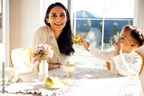 Woman with long dark hair smiles joyfully at a table while a young boy playfully offers her an apple, surrounded by flowers and a cozy, bright kitchen atmosphere