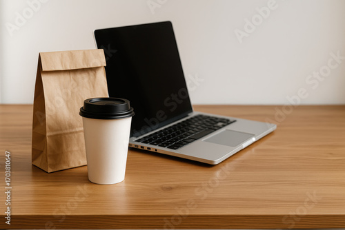 Work-life balance lunch with laptop and coffee: A laptop, takeaway coffee cup and a brown paper bag representing lunch or breakfast, sitting on a wooden desk with bright light.