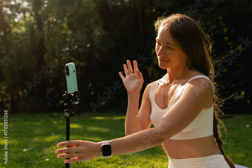 Young woman in sportswear stretching body on fitness mat at public park. Beauty blogger filming yoga in morning park. Vlogger live streaming. User-generated content UGC. Part of a series. Soft focus