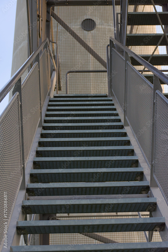 Fototapeta premium A vertical, low-angle shot of a wide metal staircase ascending toward a platform, emphasizing its repetitive pattern and industrial design.