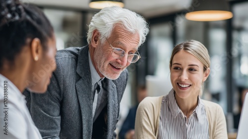 A group of business professionals communicates in a modern office environment. An older man shares insights with two women, fostering collaboration and teamwork during a workday.