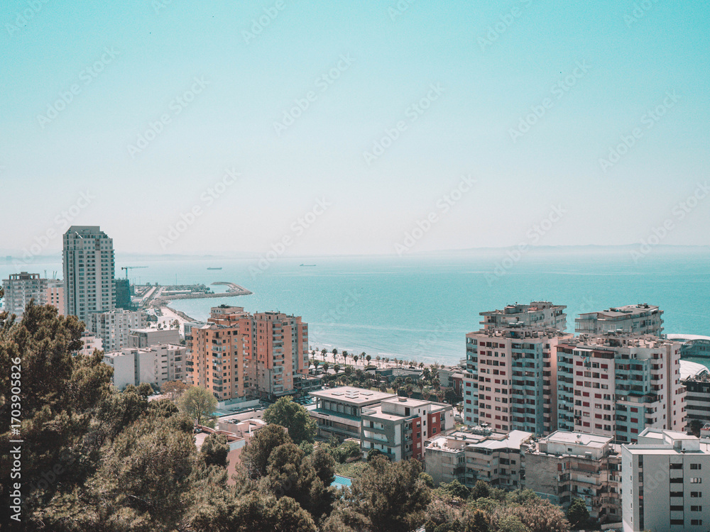 Obraz premium Stunning coastal cityscape photo of modern buildings along a turquoise shoreline, framed by lush greenery under a clear sky. Ideal for travel decor, seaside art, and eco-friendly designs.