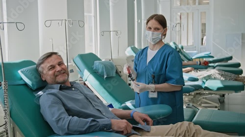 Portrait shot of masked nurse in blue scrubs and smiling middle-aged man posing for camera after donating blood in clinic