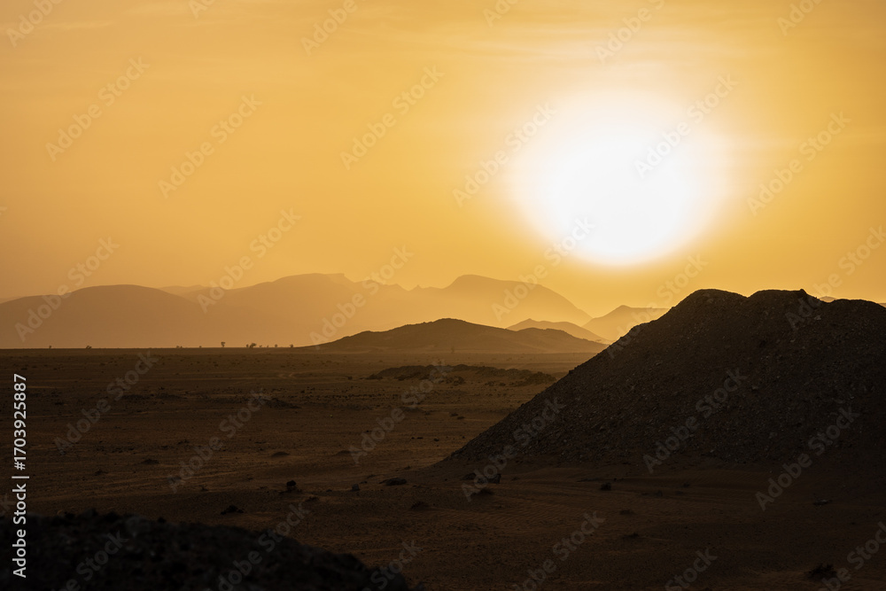 Obraz premium Golden Sahara desert sunset in Morocco with bright sun over distant mountain silhouettes and dark sand dunes creating dramatic evening landscape view