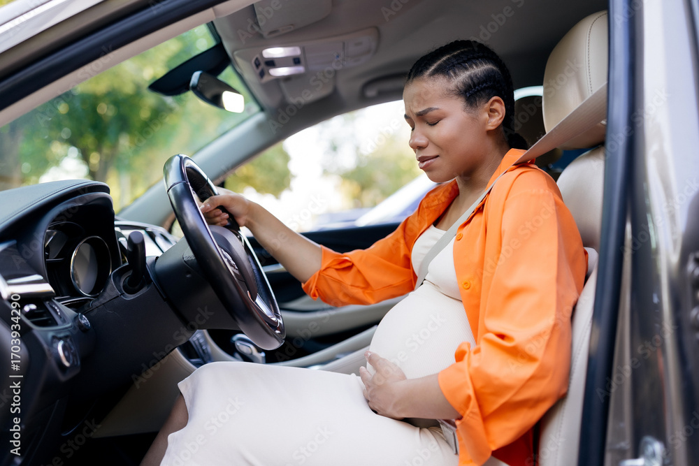 Obraz premium Worried young African American pregnant woman sitting in car, hand on belly, feeling pain or contraction. Concept of maternity stress and urgent health concern