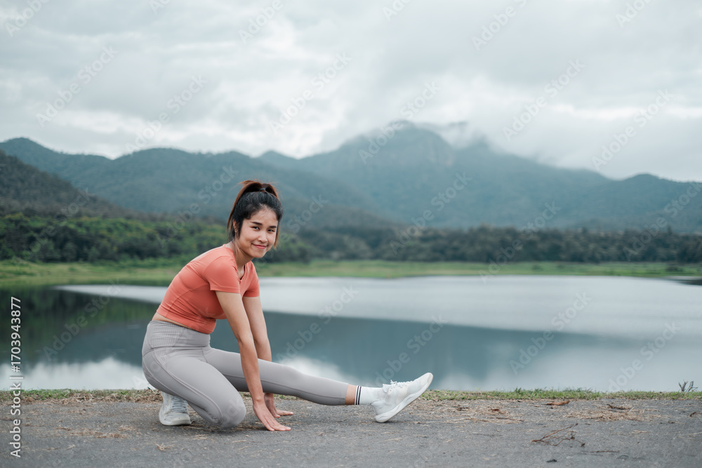 Fototapeta premium Woman in activewear stretching by a lake with mountains in the background, showcasing fitness and nature.
