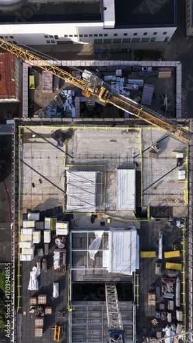 Drone view of an active construction site with a yellow crane, concrete structures, and building materials organized on the ground.