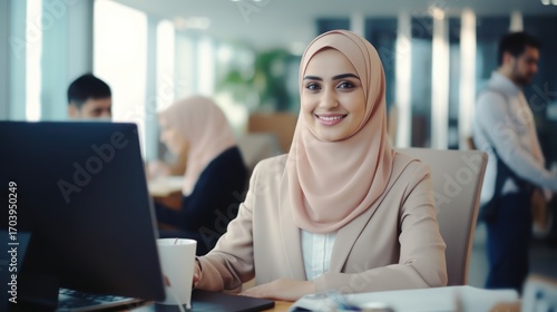 A young woman wearing a hijab sits at a desk in an office, smiling at the camera.
