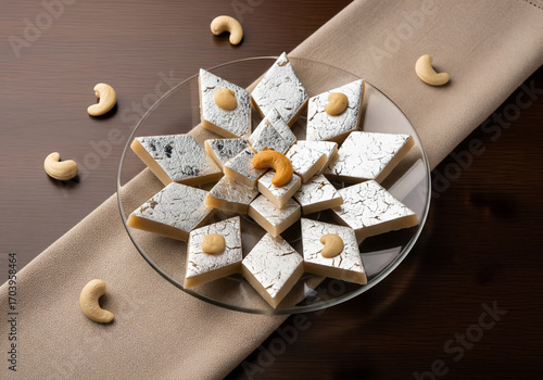 A plate of traditional Indian cashew nut barfi, known as kaju katli, decorated with edible silver foil and cashews on a brown background.