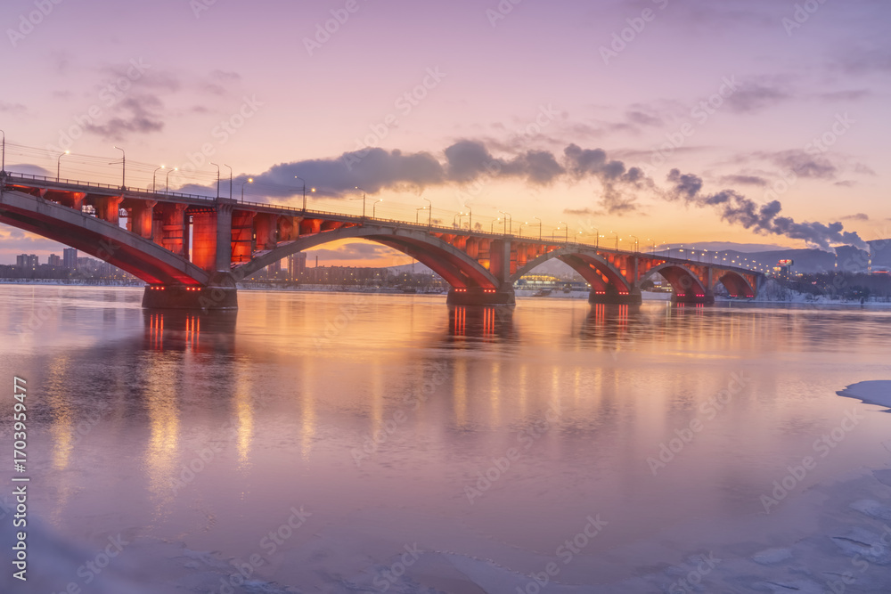 Naklejka premium Russia is an old bridge in the center of Krasnoyarsk across the Siberian Yenisei River, known as the Communal Bridge connecting the left and right banks of the city at the hour of colorful pink dawn.