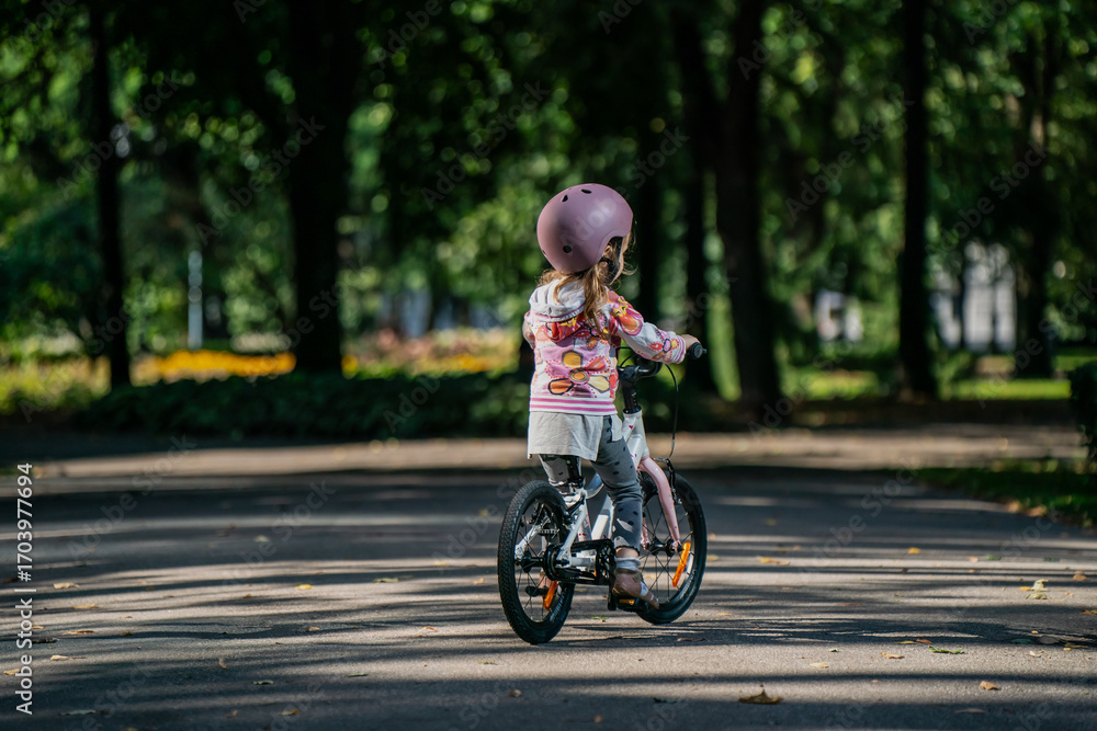 Obraz premium Young child wearing pink helmet and colorful hoodie riding small bicycle along park pathway, practicing balance and cycling skills outdoors in safe green environment surrounded by trees