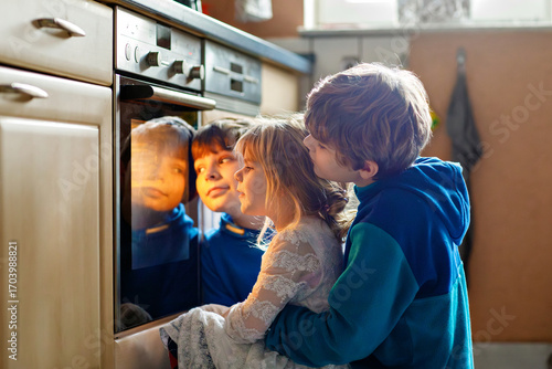Little toddler girl and two kids boys baking pumpkin cake or cookies in kitchen. Three children, siblings, brothers and sister sitting near oven and waiting. On thanksgiving or christmas holiday.
