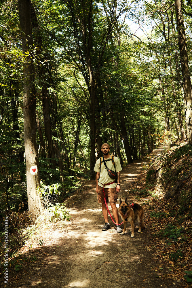 Fototapeta premium Man hiking with German Shepherd on forest trail in Fruska Gora National Park, Serbia. Front view portrait. Hiking with pet concept.