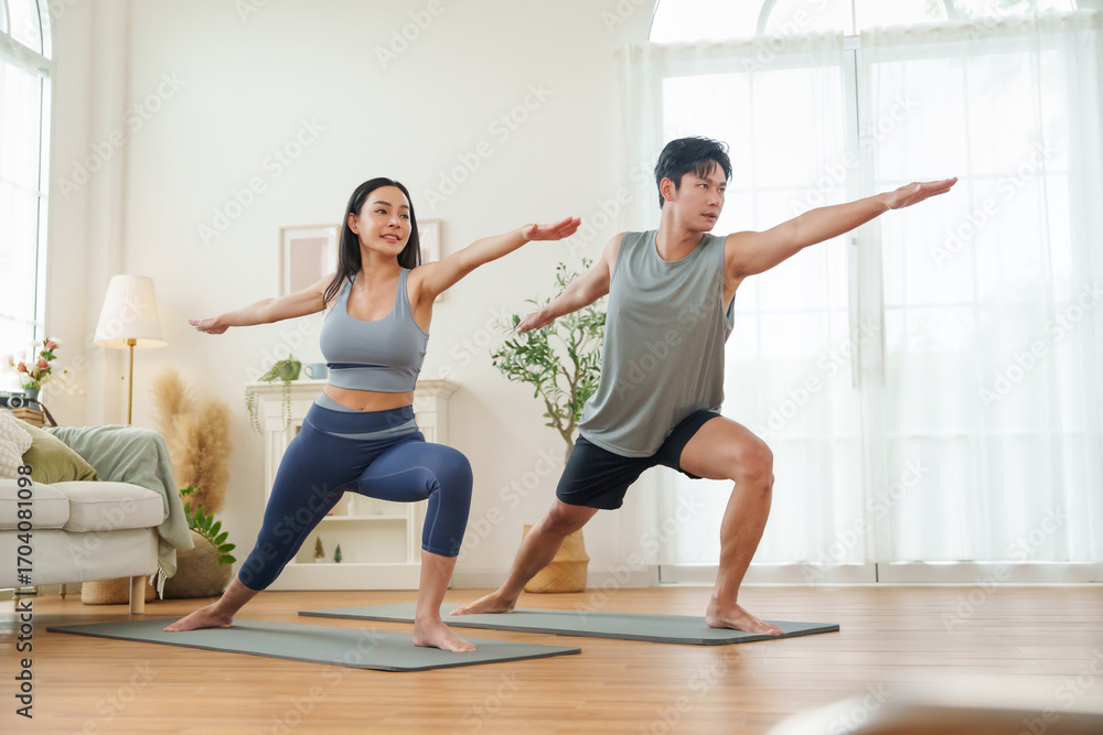 Fototapeta premium Asian couple stands strong in Warrior II pose on yoga mats, focusing together in a cozy living room.