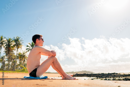 Quadro su tela Young man in swim briefs sitting on the beach, relaxing under the sun with palm trees in the background