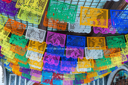A colorful display of paper lanterns hangs from the ceiling. The lanterns are of various colors and sizes. Day of the Dead celebration in Mexico, papel picado crafts, offerings, La Catrina