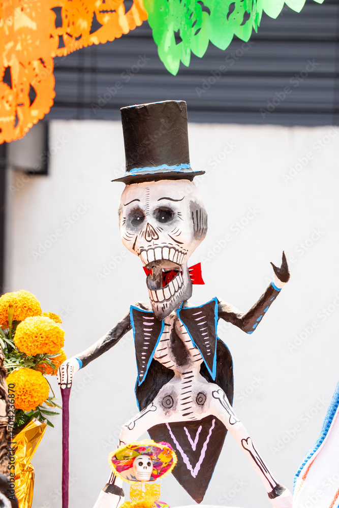 Fototapeta premium A skeleton dressed in a suit and top hat is holding a flower. The skeleton is smiling and he is happy. Day of the Dead celebration in Mexico, papel picado crafts, offerings, La Catrina